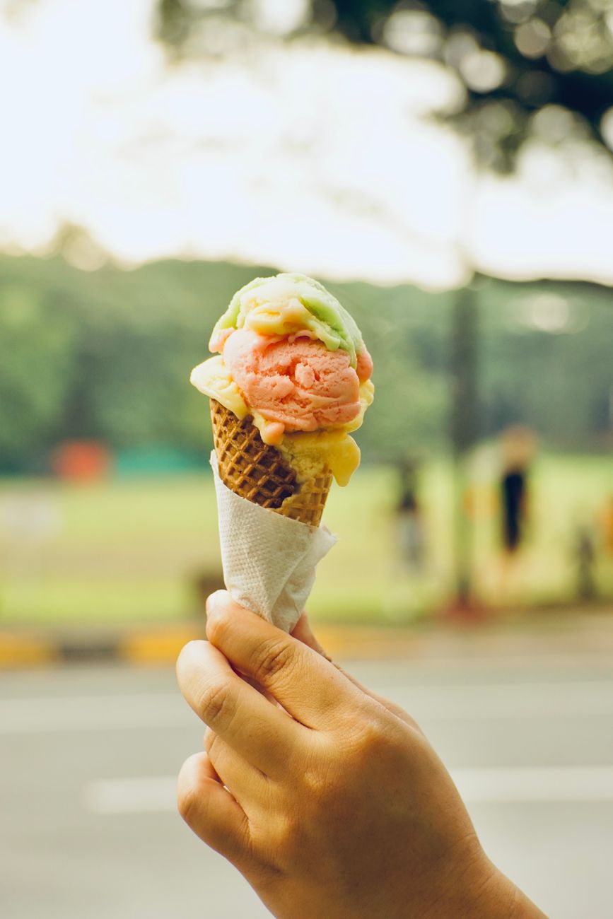 A hand holds a vibrant ice cream cone outdoors in a sunny park scene.
