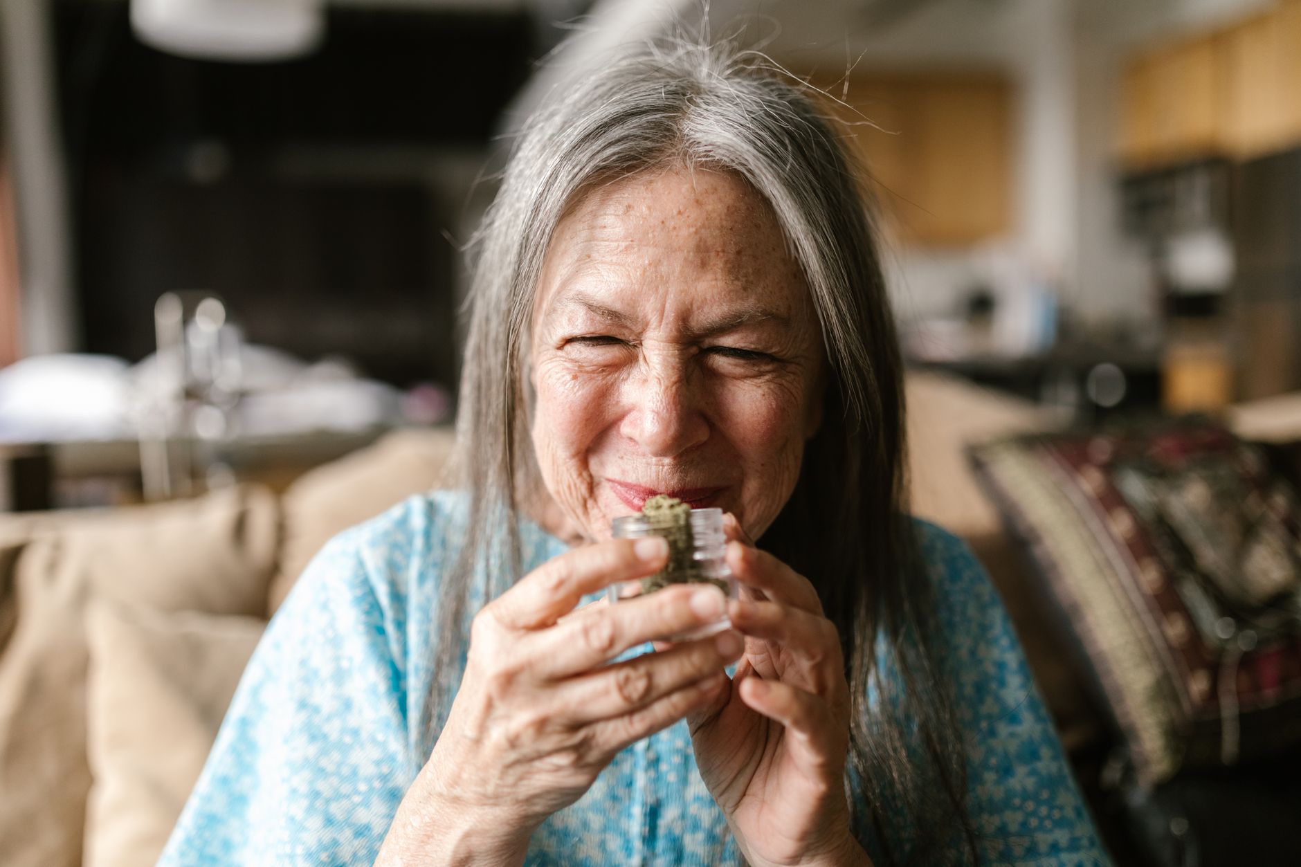 Senior woman enjoying the aroma of cannabis, smiling warmly in a cozy indoor setting.